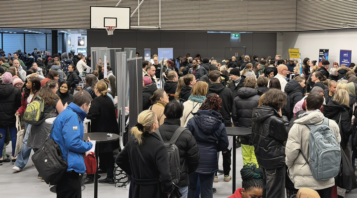 Sisätiloissa järjestetty tapahtuma, jossa suuri joukko ihmisiä seisoo tiiviisti esittelypisteiden ja roll-upien ympärillä; osallistujilla on talvivaatteet ja reppuja, ja tila on liikuntasali.