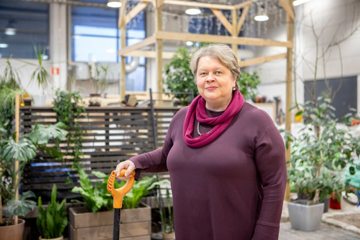 Mirja Hannula katsoo kameraan. Hän pitää kädessään puutarhalapiota. Taustalla näkyy kasveja ja puurakennelma.