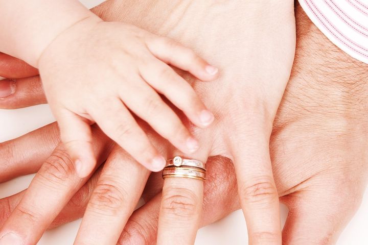 Kolme kättä päällekkäin: vauvan käsi, aikuisen käsi vihkisormuksilla ja vielä yksi aikuinen käsi.