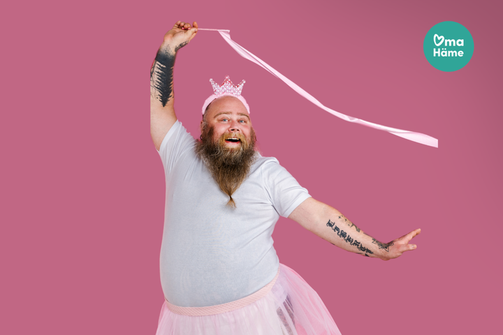 Aikuinen mies hymyilee, pukeutunut vaaleanpunaiseen tyllihameeseen ja kruunuun, heiluttaen vaaleanpunaista nauhaa.