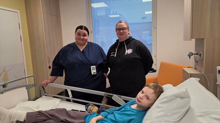 Nuori poika makaa sairaalasängyssä, ja hänen vierellään seisovat kaksi aikuista henkilöä sairaalan huoneessa.