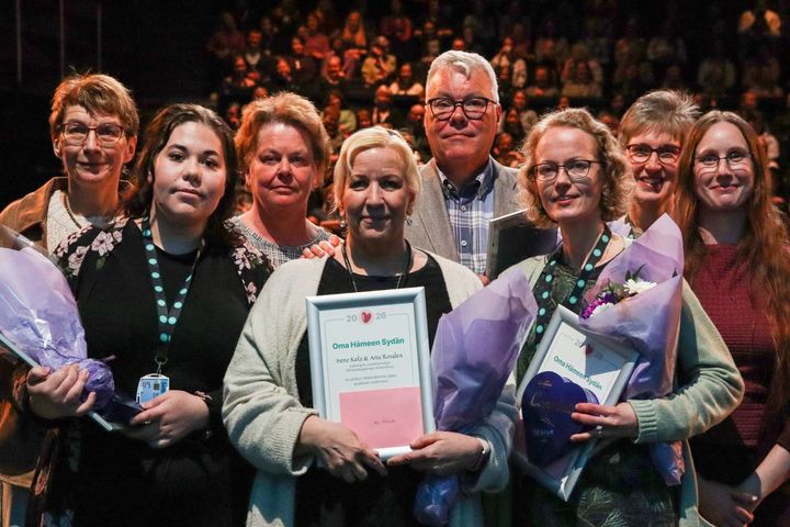 Ryhm%E4%20ihmisi%E4%20seisoo%20lavalla%2C%20joillakin%20on%20kukkakimppuja%20ja%20papereita%20k%E4dess%E4%E4n.%20Taustalla%20yleis%F6.