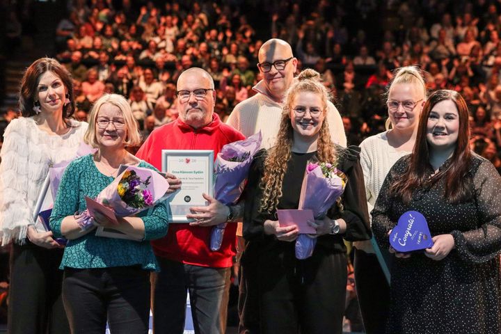 Ryhmä ihmisiä seisoo lavalla, joillakin on kukkakimppuja ja papereita kädessään. Taustalla yleisö.