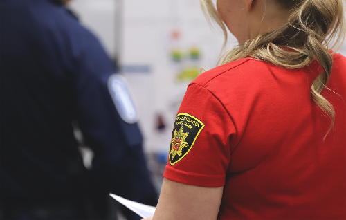 Nainen punaisessa paidassa, jossa on pelastuslaitoksen logo, seisoo poliisin kanssa.