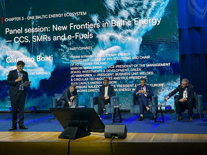 Paneelikeskustelu energiamessuilla, viisi henkilöä lavalla, taustalla aaltojen kuva ja esityskehotteet.