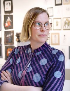 Henna Ryynänen seisoo raitakuosisessa mekossa käsivarret ristissä. Hän katsee oikealle. Hänellä on vaaleat polkkamittaiset hiukset. Taustalla näkyy taideteoksia seinällä.