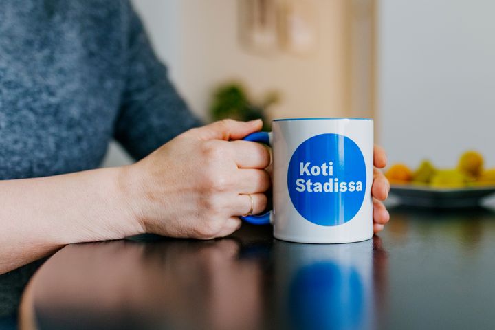 Kädet ja muki, jossa Hason slogan Koti Stadissa