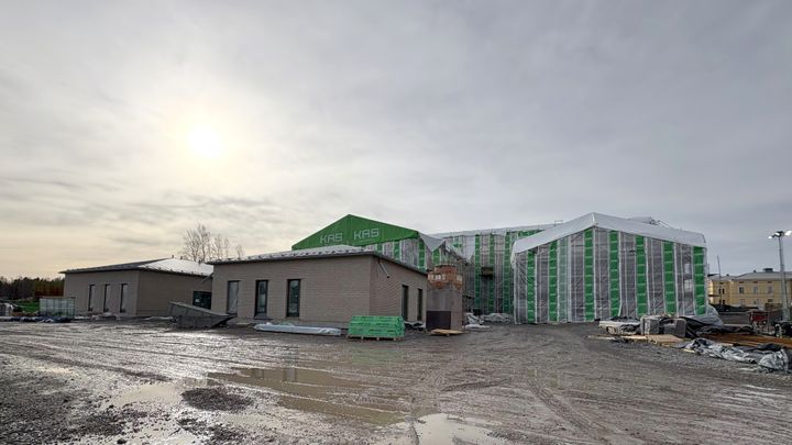 Rakennustyömaa, jossa on keskeneräinen suuri rakennus peitettynä säänsuojalla. Edessä pienempiä, jo valmiimpia rakennuksia. Taustalla pilvinen taivas ja aurinko.