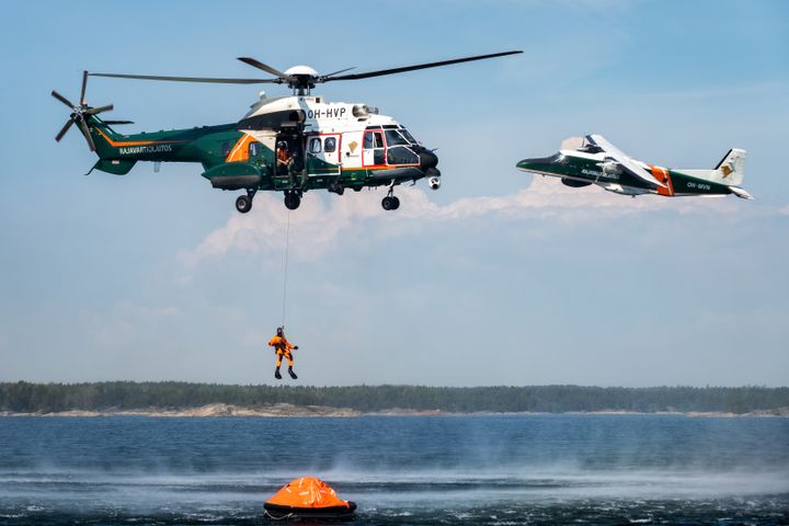 Helikopteri nostaa pelastustyöntekijää vedestä pelastuslautalla. Toinen lentokone lentää taustalla.