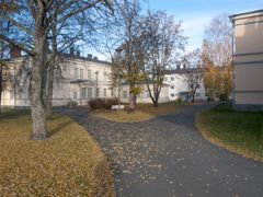 Syksyinen puistomaisema, jossa on vanha vaalea rakennus, kävelytie ja pudonneita lehtiä puiden alla.