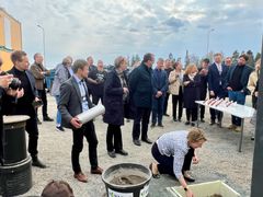 Joukko ihmisiä ulkona rakennustyömaalla, yksi henkilö heittää laastia peruskiveen.