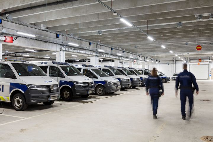 Rivi poliisiautoja pysäköitynä autotallissa, kaksi ihmistä kävelee poispäin.