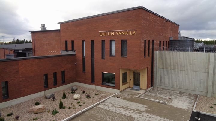 Punatiilinen rakennus, jossa lukee "Oulun vankila". Ympärillä on piha-alue kivillä ja kasveilla.