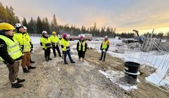 Rakennustyömaa, jossa kypäriä ja huomioliivejä käyttäviä henkilöitä seisoo peruskivenmuuraustilaisuudessa. Taustalla rakennustyökoneita ja puita.