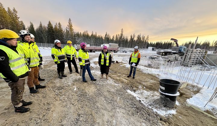Rakennustyömaa, jossa kypäriä ja huomioliivejä käyttäviä henkilöitä seisoo peruskivenmuuraustilaisuudessa. Taustalla rakennustyökoneita ja puita.