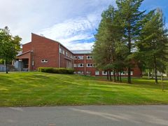 Punatiilinen rakennus vehreän nurmikon ja puiden keskellä. Kirkas taivas taustalla.