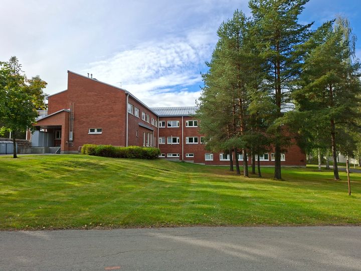 Punatiilinen rakennus vehreän nurmikon ja puiden keskellä. Kirkas taivas taustalla.