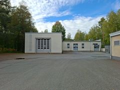 Vaalea rakennus, jossa on nosto-ovi ja kaksi suurta ovea. Edustalla on asfalttipiha ja taustalla vihreää metsää.