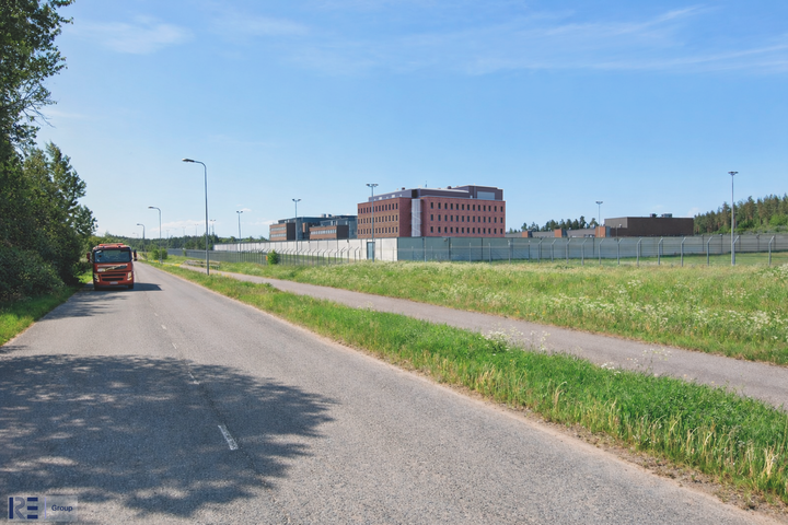 Maantie, jonka oikealla puolella korkean aidan ympäröimä tiilirakennuskompleksi, nurmikkoa ja valaisinpylväitä kirkkaan sinisen taivaan alla.