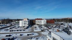 Ilmakuva kahdesta valkoisesta monikerroksisesta rakennuksesta ja ruskeasta rakennusosasta, edessä pysäköintialueita ja katu lumisessa ympäristössä.