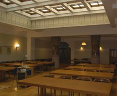 Suuri sali, jossa on useita pitkiä puupöytiä ja tuoleja, katossa koristeellisia lasi-ikkunoita.