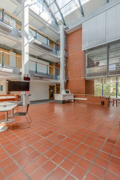 Avara sisätila, jossa on punatiililattia, korkeat lasiseinät ja katto. Oikealla puolella on punatiiliseinä ja vasemmalla olevassa pöydässä on tuolit. Paljon luonnonvaloa tulvii sisään lasikaton kautta.