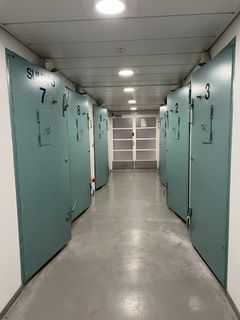 Kapea käytävä, jonka molemmin puolin on metallisia ovia, numeroituina 7-12 vasemmalla ja 2-6 oikealla.