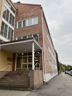 Kolmikerroksinen punatiilinen rakennus, jossa on valkoinen katos ja portaat sisäänkäynnille. Pilvinen taivas ja puiden reunustama katu.