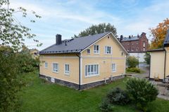 Keltainen puutalo vehreällä pihalla, taustalla näkyy punatiilinen kerrostalo.