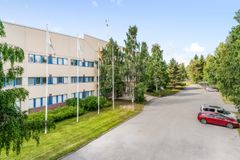 Kolmikerroksinen vaalea rakennus, jonka edessä on lipputankoja ja pysäköityjä autoja. Rakennusta ympäröi vehreä maisema.