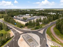 Ylhäältä kuvattu moderni liikenneympyrä vehreässä ympäristössä, taustalla rakennuksia ja metsää.