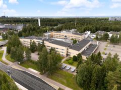 Ilmakuva laajasta vaaleasta rakennuksesta metsän keskellä, jonka ympärillä on parkkialue.