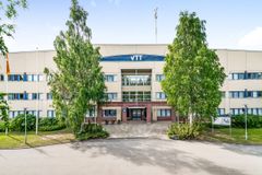 Vaalea rakennus, jonka keskellä lukee "VTT". Puita kasvaa molemmin puolin.