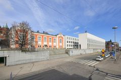 Kuva näyttää kadun, jonka vasemmalla puolella on oranssi ja valkoinen rakennus. Oikealla on moderni valkoinen rakennus. Kadulla on pysäköityjä autoja ja suojatie. Taustalla on sininen taivas.