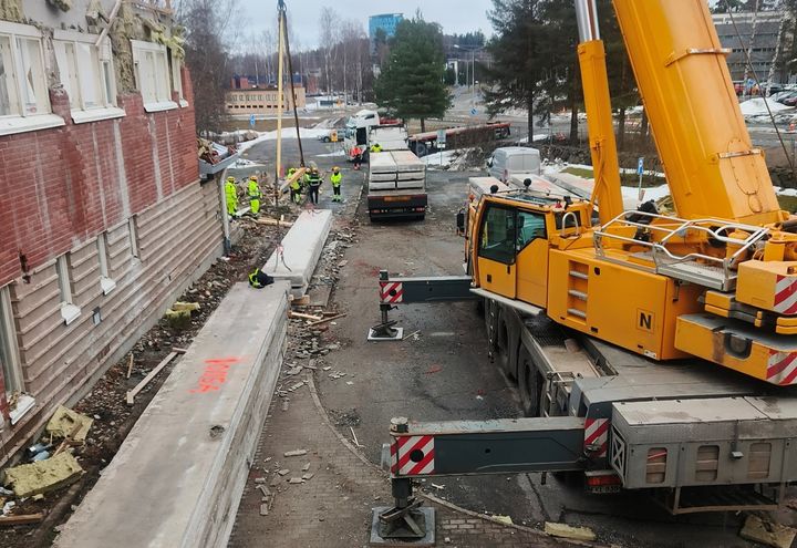 Rakennustyömaa, jossa suuri keltainen nosturi kuormaa betonilevyjä kuorma-autoon. Työmiehiä ja purkurakennus taustalla.
