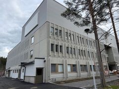 Vaalea kolmikerroksinen rakennus, jossa suuret ikkunarivit. Rakennuksen ympärillä viimeisteltävää piha-aluetta pilvisenä päivänä.