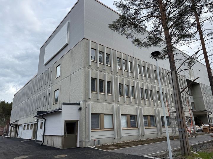 Vaalea kolmikerroksinen rakennus, jossa suuret ikkunarivit. Rakennuksen ympärillä viimeisteltävää piha-aluetta pilvisenä päivänä.