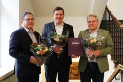 Kolme miesoletettua henkilöä puvut päällä, kukkakimput käsissään.
