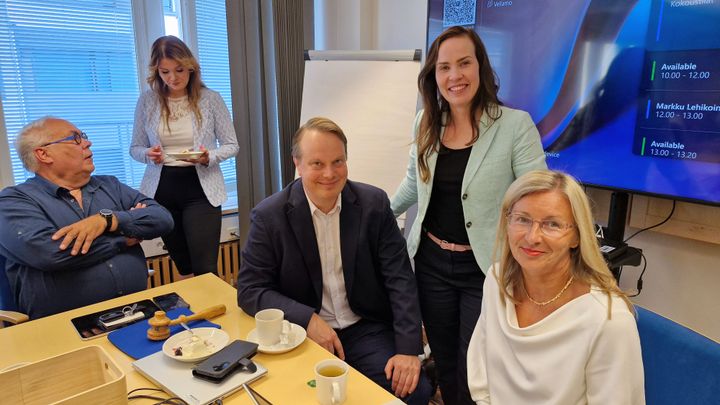 Kokouspöydän ääressä istumassa kaksi miestä ja yksi nainen. Heidän takanaan seisoo kaksi naista.