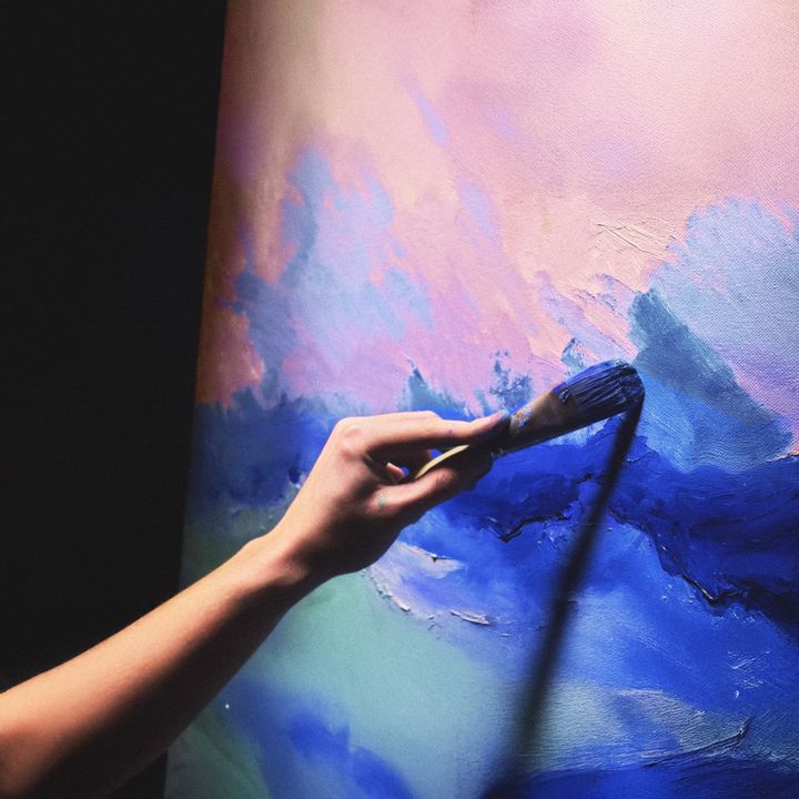Taideteosta maalaavassa kädessä sivellin, abstrakti maalaus, jossa sinisen eri sävyjä ja vaalean punaisen ja vaalean vihreän sävyjä.