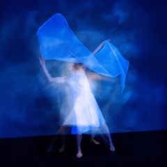 Sininen tausta, tanssija, jolla valkoinen mekko ja kädessä ohut valkoinen harso.