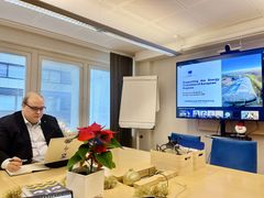 Tummaan pukuun pukeutunut mies istuu kokoustilassa pöydän ääressä. Pöydällä tietokone, muistikirjoja ja joulutähti. Taustalla iso näyttö, jossa teksti: Supporting the Energy Transition of Europea n Regions.