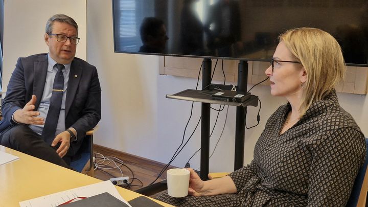 Nainen ja mies istuvat kokoustilassa pöydän ääressä rinnakkain keskustelemassa. Naisella on kädessään kahvikuppi.