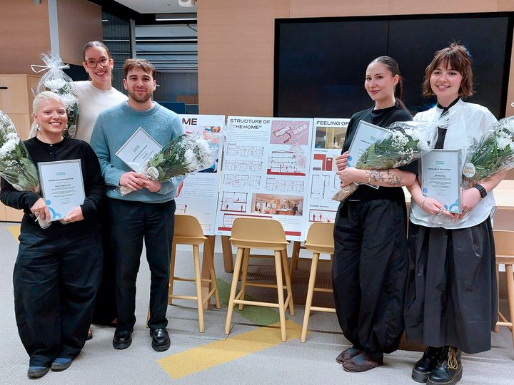 Kuvassa viisi henkilöä, jotka pitävät kukkakimppuja, sertifikaatteja ja seisovat suunnitelmien edessä.