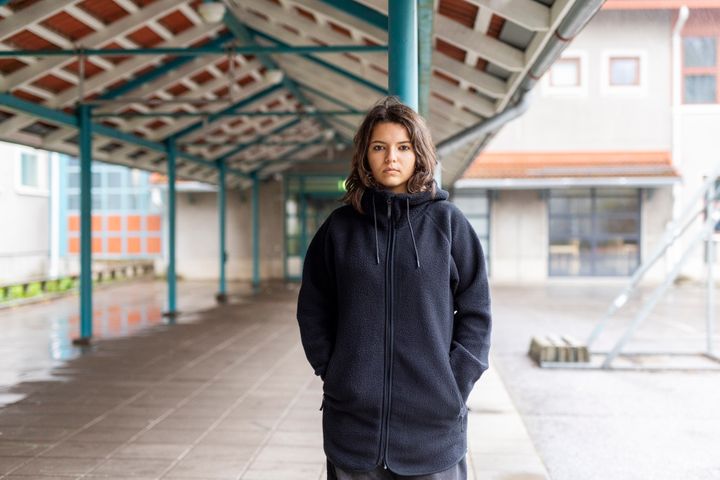 Tyttö nojaa ulkona pylvääseen ja katsoo suoraan kameraan vakavana. Taustalla asfalttia ja katos