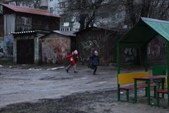 Kaksi lasta juoksee leikkikentällä talvisessa ympäristössä. Taustalla näkyy rapistuneita rakennuksia.