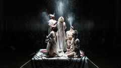 Viisi henkilöä seisoo ja polvistuu valaistulla lavalla, jota ympäröi pimeys.