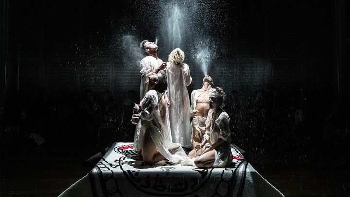 Viisi henkilöä seisoo ja polvistuu valaistulla lavalla, jota ympäröi pimeys.