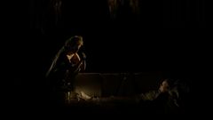 Näyttämökohtaus, jossa tummaan pukeutunut henkilö nojaa eteenpäin puhuen sohvalla makaavalle henkilölle hämärässä valaistuksessa.