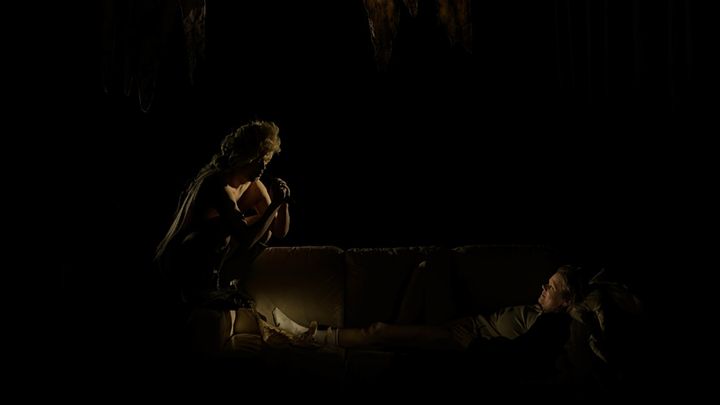Näyttämökohtaus, jossa tummaan pukeutunut henkilö nojaa eteenpäin puhuen sohvalla makaavalle henkilölle hämärässä valaistuksessa.
